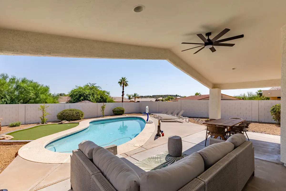 This image captures a luxurious backyard setup featuring a swimming pool, an outdoor seating area with comfortable chairs, well-tended landscaping, and palm trees framing the scene. The setting is tranquil yet inviting, perfect for both daytime relaxation and evening gatherings.
