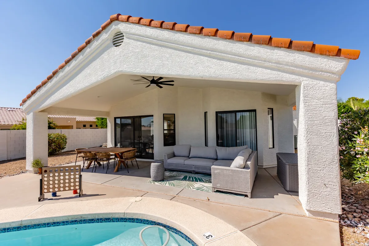 A sun-drenched backyard featuring a sparkling swimming pool, ample outdoor seating, and a dining table set beneath a shaded patio. The area is surrounded by lush landscaping and designed for relaxation, outdoor meals, and family fun.