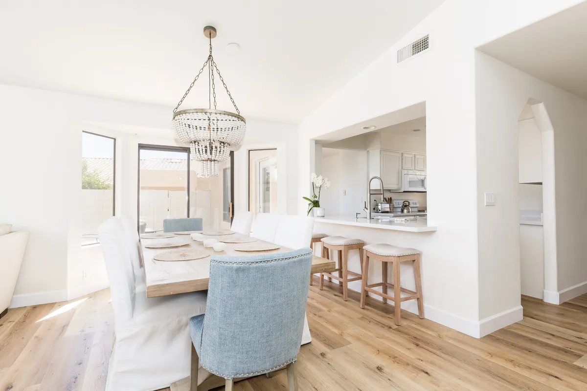 A warm and inviting dining area with a stylish chandelier over a large dining table, adjacent bar stools, and a convenient kitchen pass-through. The setting blends formal dining with casual gathering, highlighting the seamless flow between spaces.