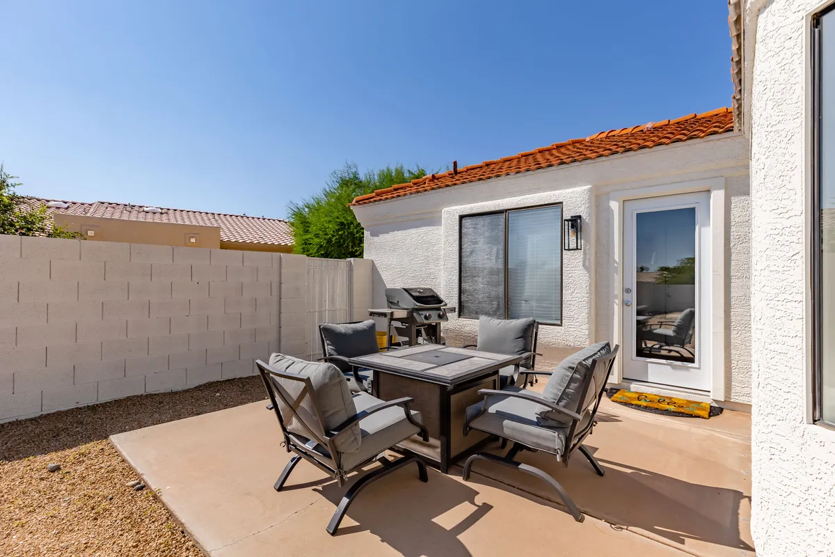 An exterior shot focused on an outdoor dining set with a grill nearby, surrounded by lush flowering plants and set against the house. The setting is casual and designed for socializing, ideal for group BBQs or enjoying meals in the fresh air.