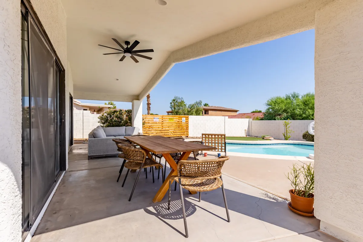 A serene backyard scene featuring an inviting swimming pool, a dining table set for two, and a cozy lounge area with potted plants. The area is bathed in natural light, suggesting either a tranquil morning or a romantic evening by the pool.
