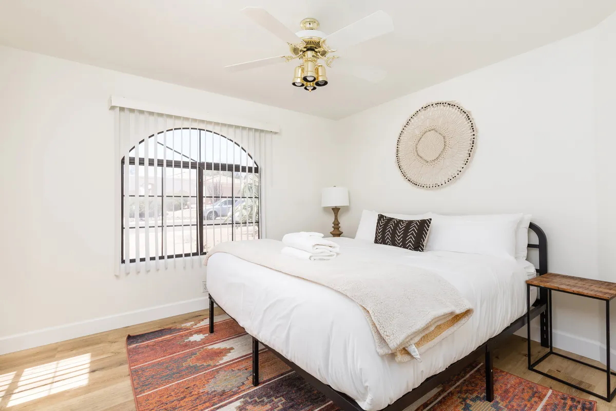 A cozy bedroom featuring a bed with white linens, a decorative wall hanging, a colorful rug, a table lamp, and sunlight streaming through the window. The intimate, inviting atmosphere suggests a restful retreat for both children and adults. There's ample space for an air mattress or baby gear add-ons.