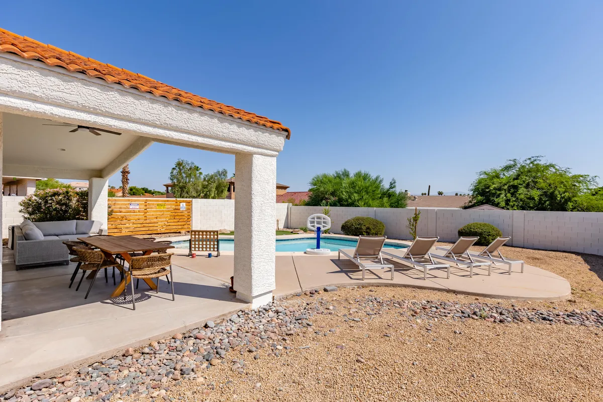 A beautifully landscaped backyard featuring a sparkling swimming pool, an outdoor dining area under a covered patio, and a set of comfortable lounge chairs. The space is inviting for both relaxation and group gatherings, with plenty of room for sunbathing, al fresco meals, or casual conversation. The warm lighting suggests it's either early evening or morning, highlighting the tranquil yet social atmosphere.